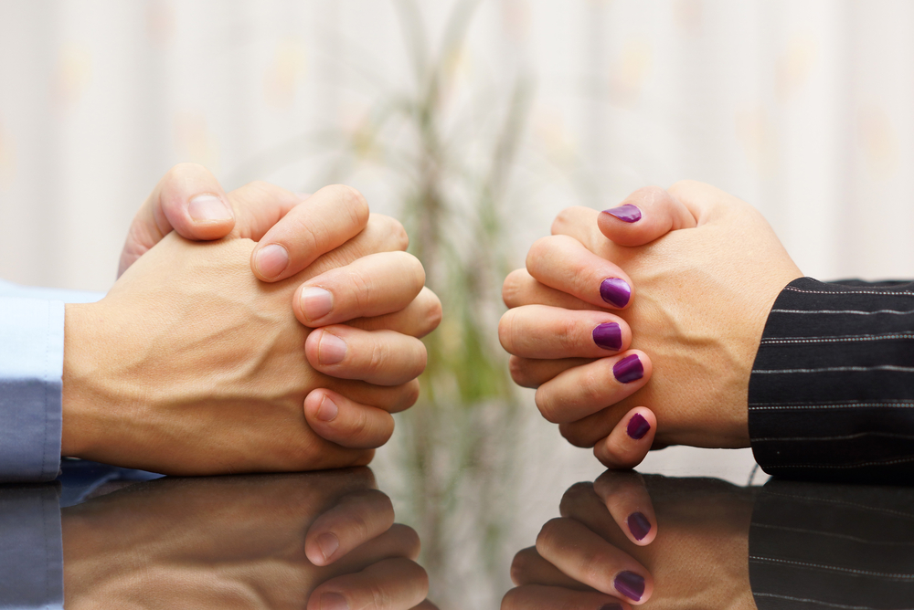 Picture of a man's hands clasped together and a woman's hands clasped together across from each other on a table.